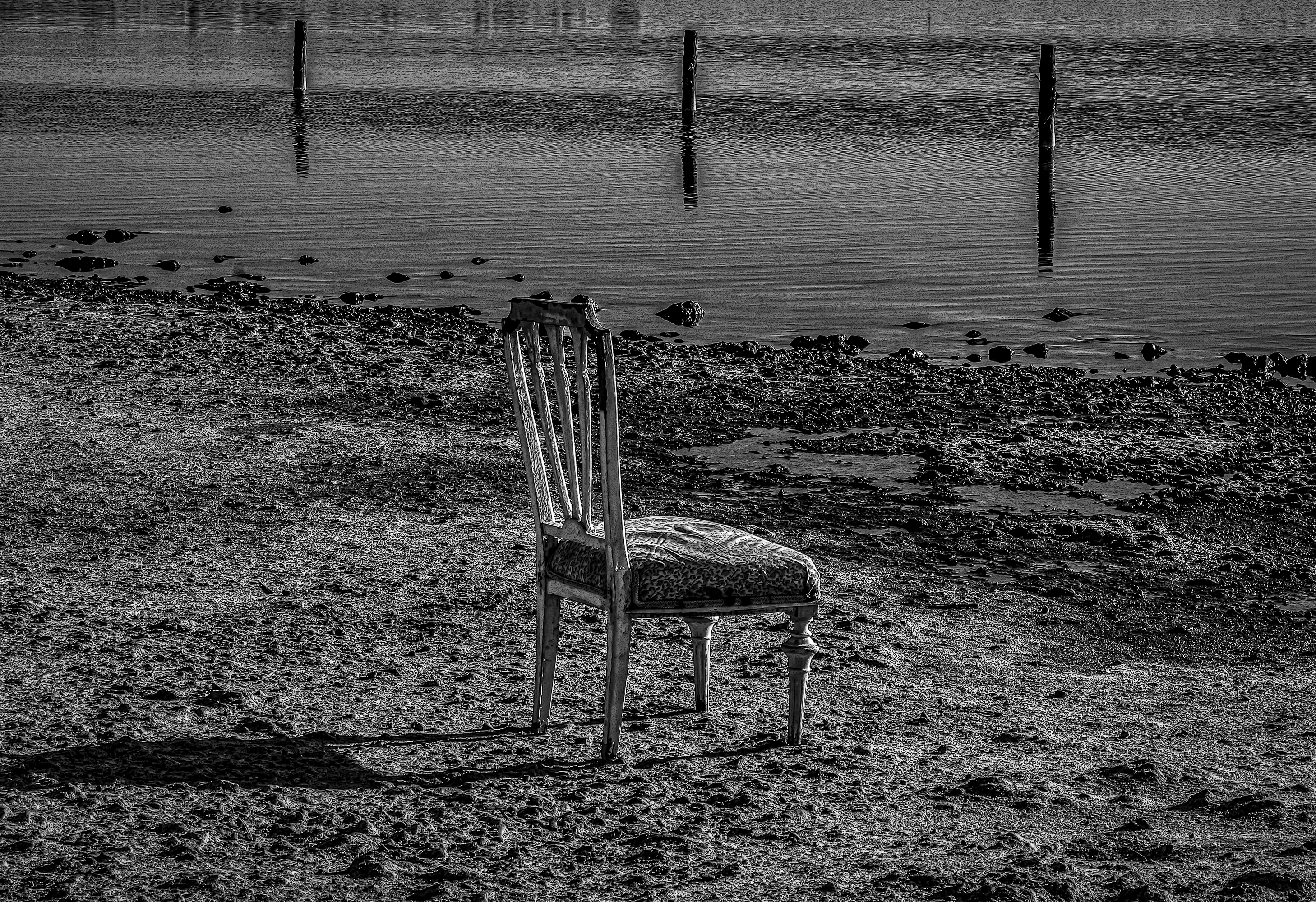 Silla de comedor solitaria en la orilla, mirando el agua con tres postes verticales al fondo