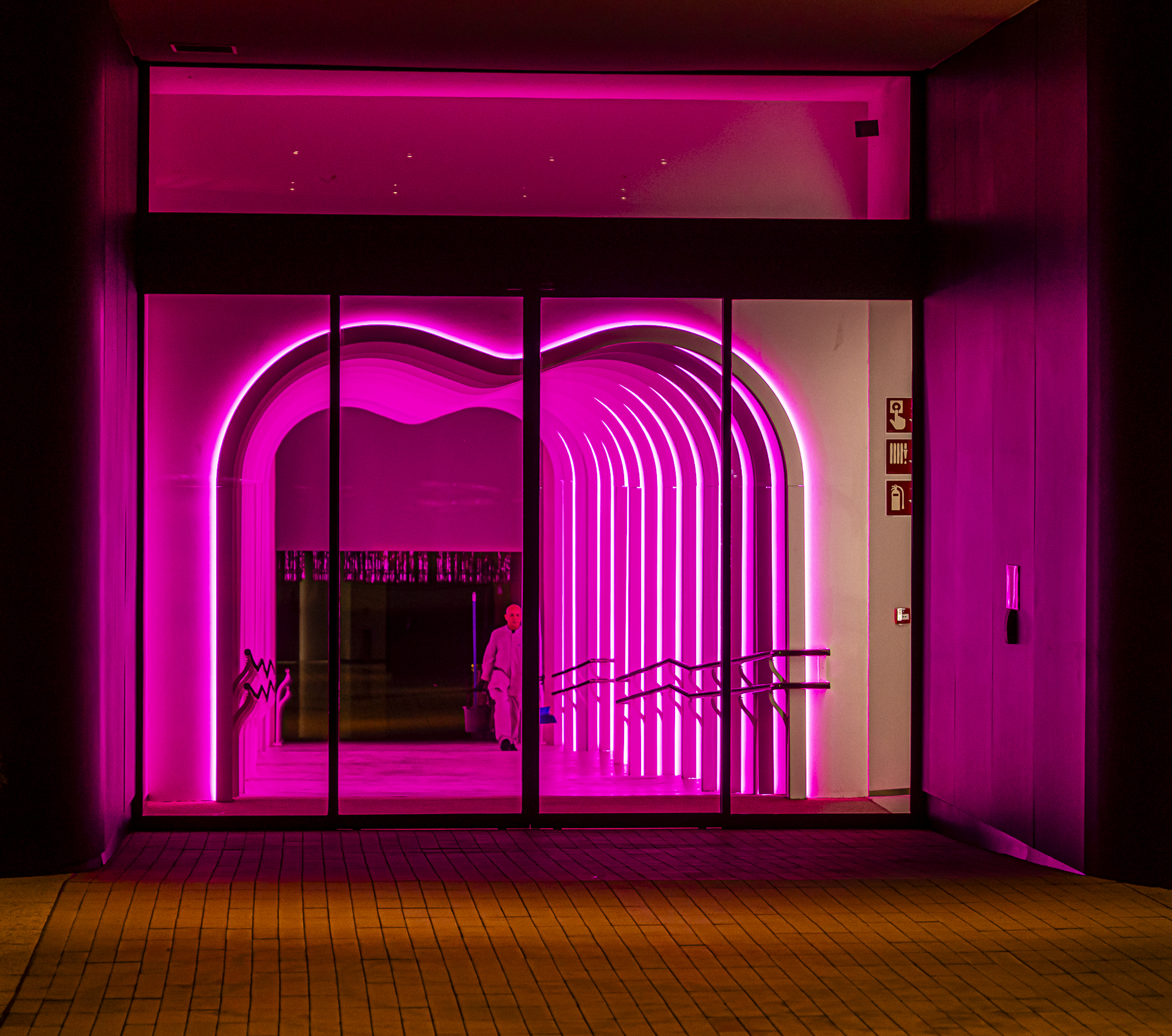 Entrada bañada en neón magenta, arcos luminosos, una persona diminuta limpiando